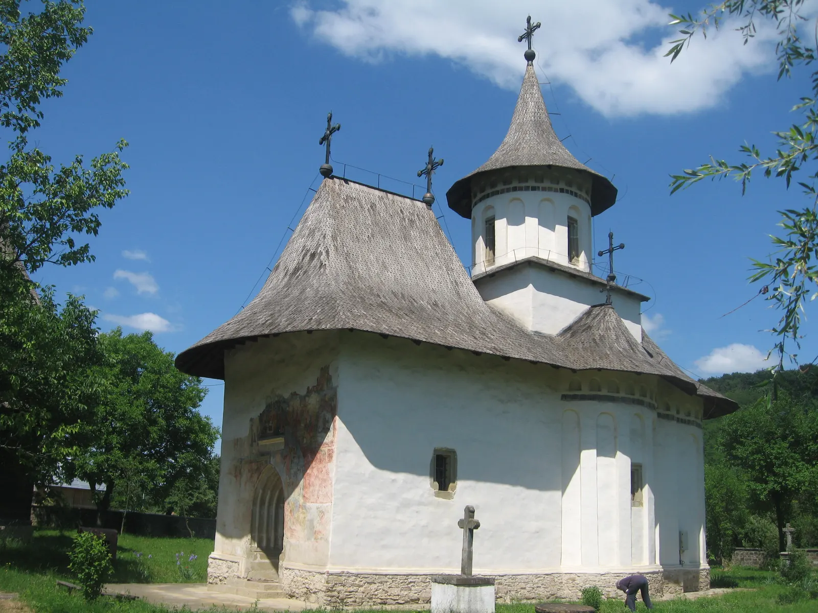Biserica din Pătrăuți, mică și albă, cu acoperiș înalt de șindrilă și turlă, iar lângă ușa de intrare, urme decolorate de pictură exterioară