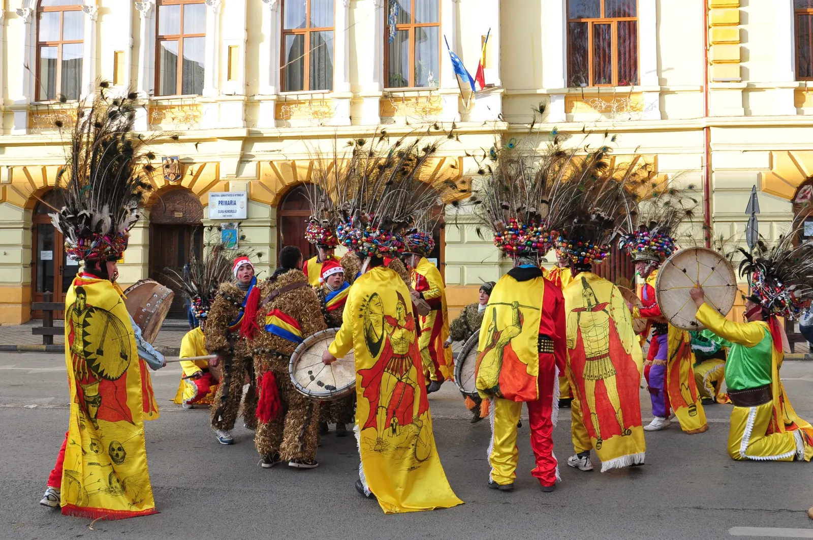 Ceată de urători cu pălării înalte din pene de păun, mantii galbene pictate și tobe mari, jucând în fața Primăriei din Gura Humorului, cu mascați în costume de paie în spate