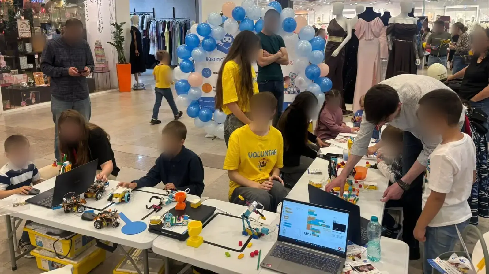 Copii la un atelier de robotică în Iulius Mall Suceava, cu roboței din piese de construcție, laptopuri și voluntari EduMax în tricouri galbene