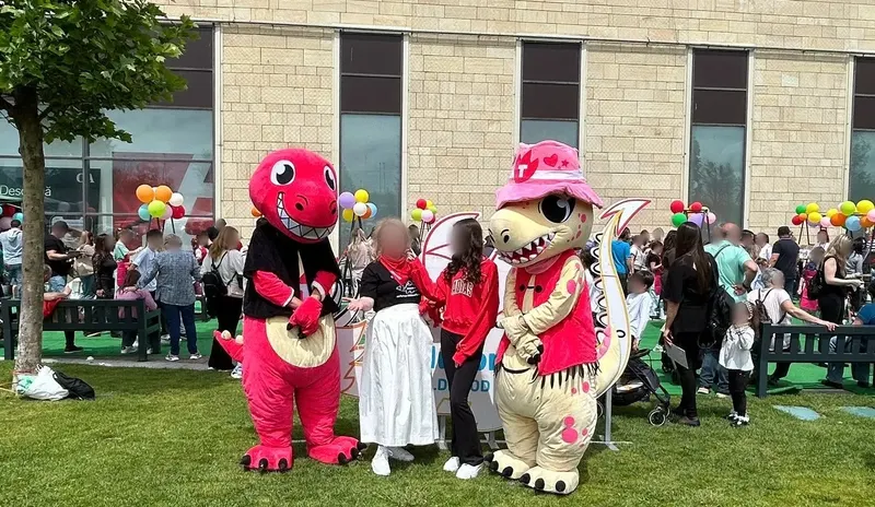 Două mascote dragon, una roșie și una crem cu pălărie roz, alături de organizatori în parcul Iulius Mall Suceava, cu baloane colorate și public în spate