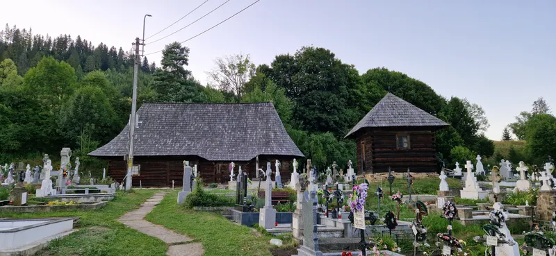 Biserica de lemn Dragoș Vodă, cu acoperiș înalt de șindrilă, și o construcție mică din bârne alături, în cimitirul satului Putna