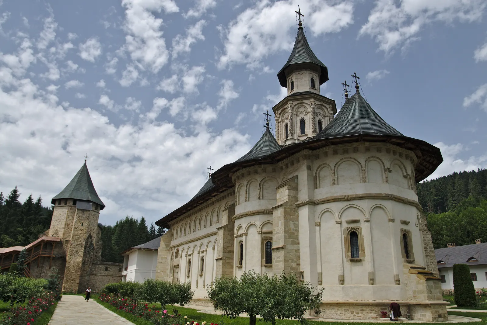 Biserica Mănăstirii Putna văzută dinspre altar, cu turla și acoperișurile întunecate, lângă Turnul tezaur din piatră și aleea cu trandafiri din incintă