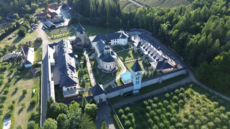 Mănăstirea Putna văzută din dronă de hramul din 15 august 2026: zidul de incintă cu turnul de poartă și clopotnița, biserica din mijloc înconjurată de oameni, Turnul tezaur în spate și pădurea din jur