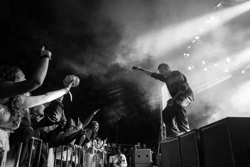 Fotografie alb-negru de la un concert de seară: solistul, urcat pe boxe, întinde mâna spre publicul de la gard