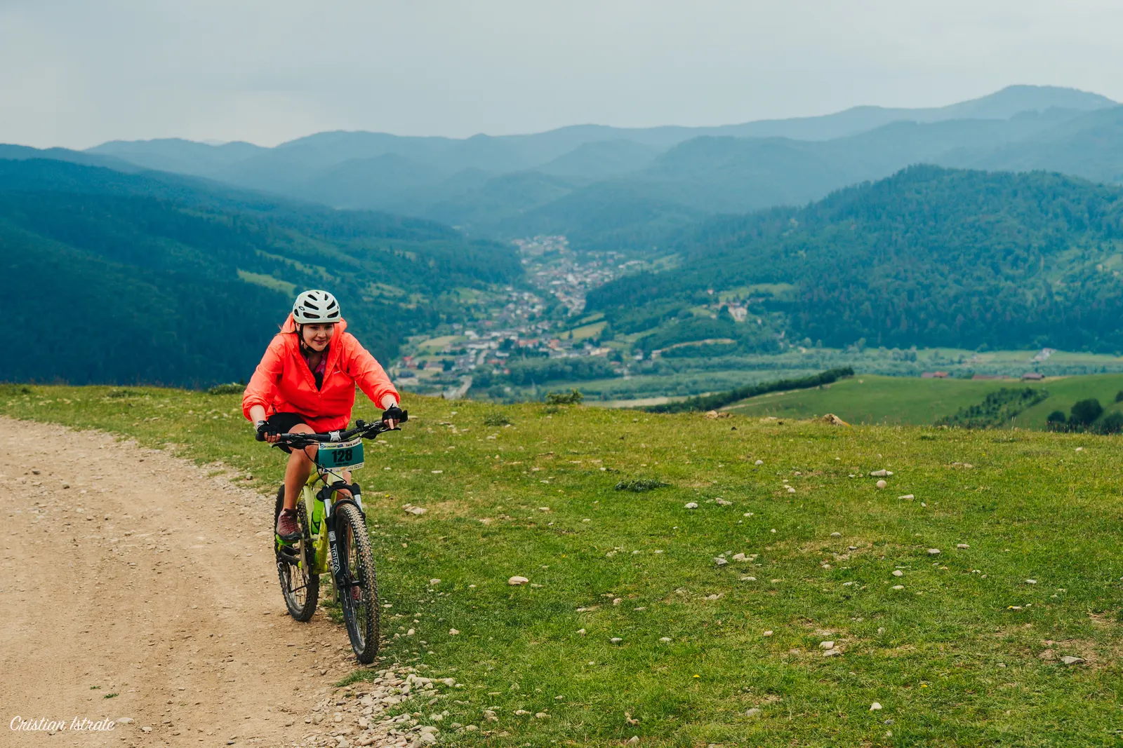 Bicicliștă cu număr de concurs urcând pe un drum de pământ pe culmea unei obcine, cu o vale locuită și dealuri împădurite în spate