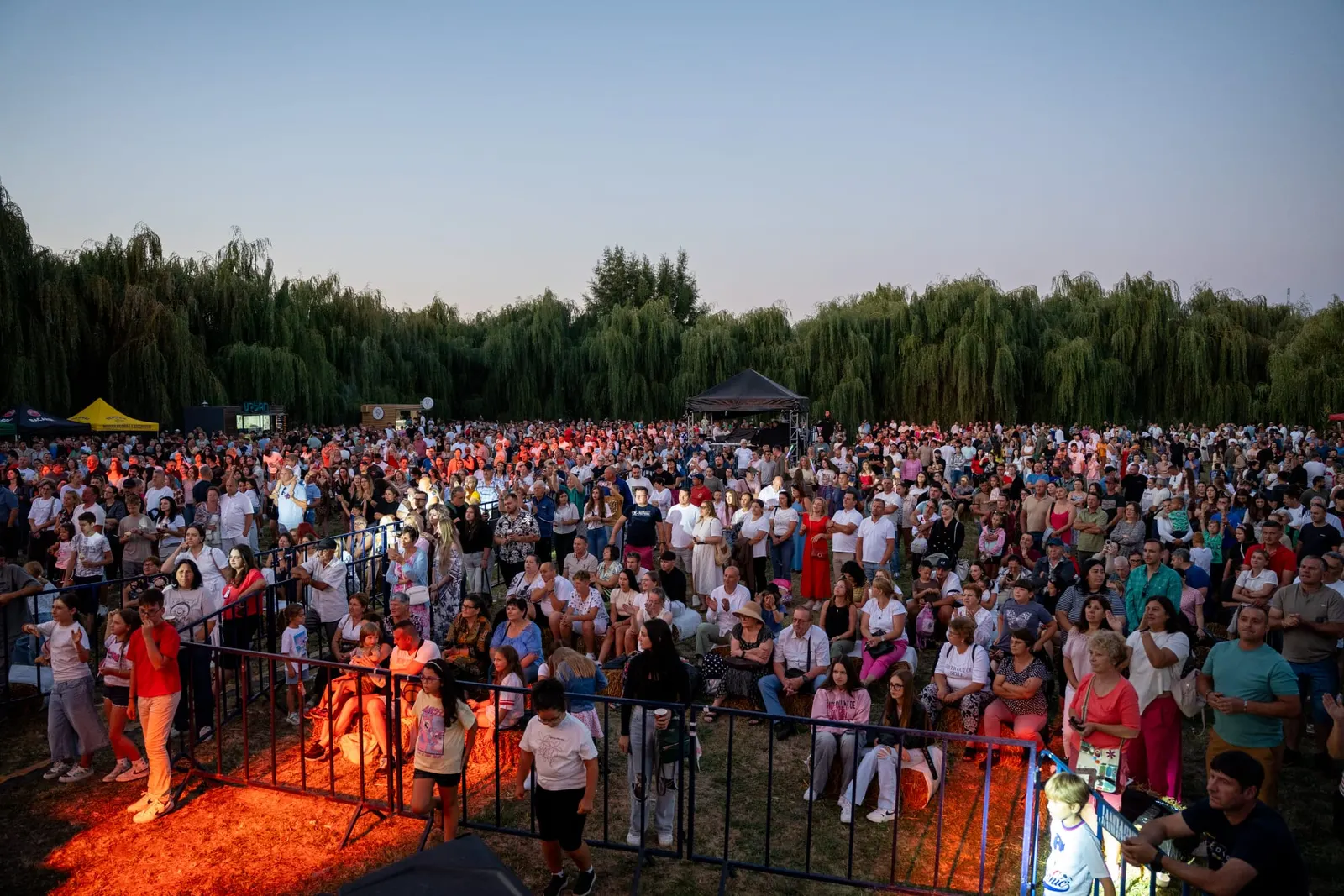 Mulțimea din fața scenei, la apus, pe pajiștea de lângă Iazul Hagigadar, cu un șir de sălcii plângătoare și standuri în fundal